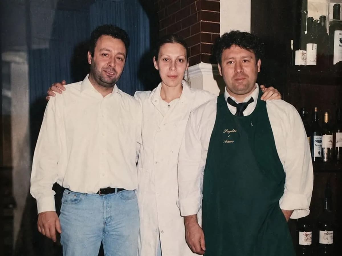 Foto storica di tre membri della famiglia in sala, davanti all'arco in mattoni e alla cantina; uno indossa il grembiule ricamato Paglia e Fieno